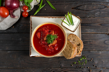Borscht with beef and sour cream. Traditional Ukrainian vegetable soup made from beets, potatoes, cereals and boiled meat, and two slices of rye bread in a ceramic bowl on a wooden kitchen table.