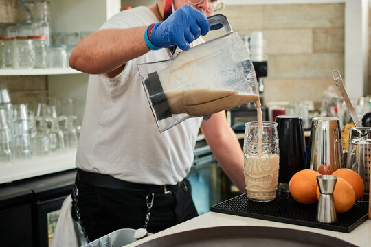 Cropped unrecognizable informal male bartender pouring milkshake in glass at counter