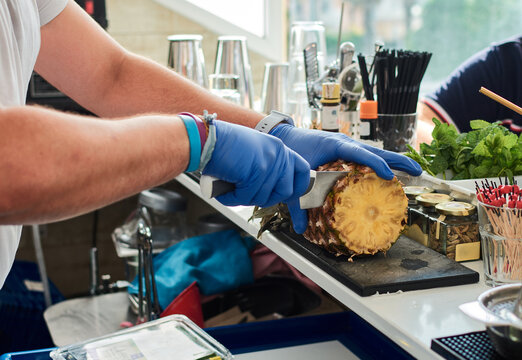 Crop anonymous male bartender in sterile gloves cutting pineapple with sharp knife while preparing healthy smoothie in summer cafe