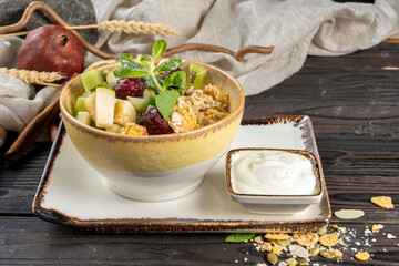 Granola with fresh fruit and yoghurt. A healthy natural breakfast of corn and oatmeal, apple, kiwi, banana, berries and yoghurt, decorated with mint leaves in a ceramic plate on a wooden kitchen table