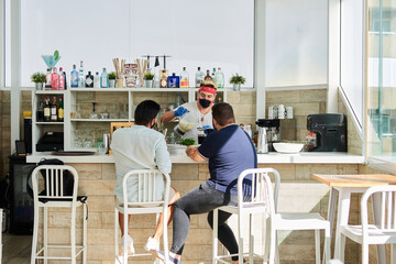 Back view of unrecognizable male clients sitting at counter in summer cafe while barman in mask and gloves preparing healthy refreshing smoothies