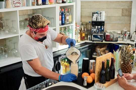 High angle of unrecognizable informal male bartender in protective mask and headband pouring milkshake in glass at counter