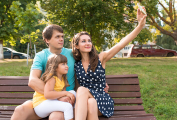 beautiful young family, woman mom, dad with daughter on her lap, take pictures of smartphone. Emotions of happiness joy fun relaxation. Summer city park. Wooden bench, green grass trees background.