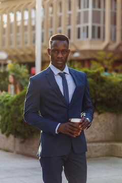 Serious adult formally dressed African American male manager drinking takeaway coffee while standing against blurred urban background of modern city