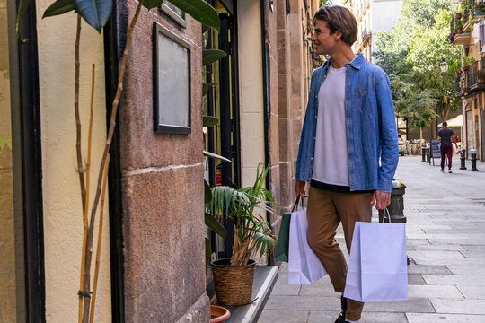 Young cheerful stylish male shopaholic in casual outfit with zero waste bags looking away in town near building