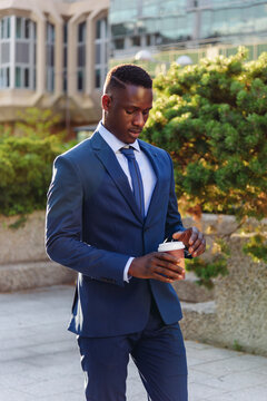 Serious adult formally dressed African American male manager drinking takeaway coffee while standing against blurred urban background of modern city