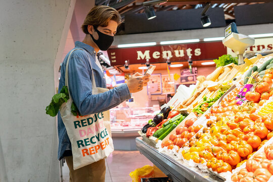 Young male customer in protective mask carrying eco friendly recycled fabric shopping bag and choosing fresh groceries while taking picture on smartphone buying food in supermarket