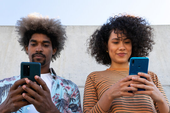 Content multiracial couple of partners in trendy clothes with Afro hairstyle standing near wall with cellphones - Powered by Adobe