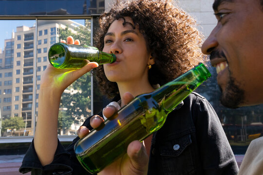 Side view of positive young African American millennial in casual clothes drinking beer while spending time together in street cafe