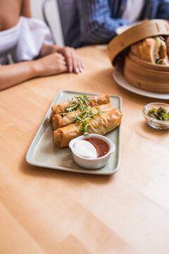 High angle of appetizing vegan stuffed spring rolls garnished with herbs served on table with sauces on wooden table in cafeteria