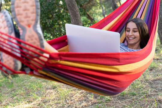 Delighted female freelancer lying in hammock and typing on netbook while working on startup project