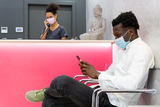 Side view of male patient in mask sitting near reception desk in modern clinic and waiting for appointment while browsing smartphone