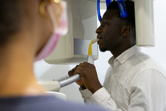 Back view of unrecognizable dentist taking X ray of teeth of black male patient sitting in medical room in modern clinic