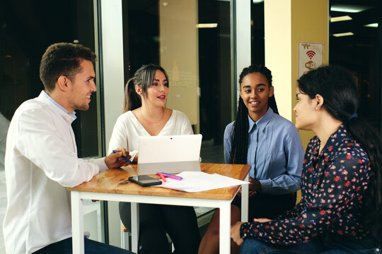 Cheerful multiethnic colleagues talking while looking at each other and sitting at table with netbook in building