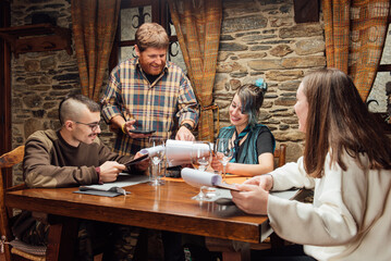 Cheerful friendly hipsters ordering food in rustic cafe while sitting together at wooden table