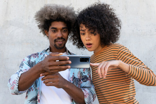 Smiling stylish black man near surprised ethnic female partner with Afro hairstyle pointing with finger at cellphone on street