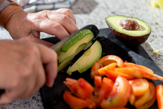 High angle crop anonymous senior female cutting ripe organic avocado and tomatoes on black board while cooking in sunny kitchen