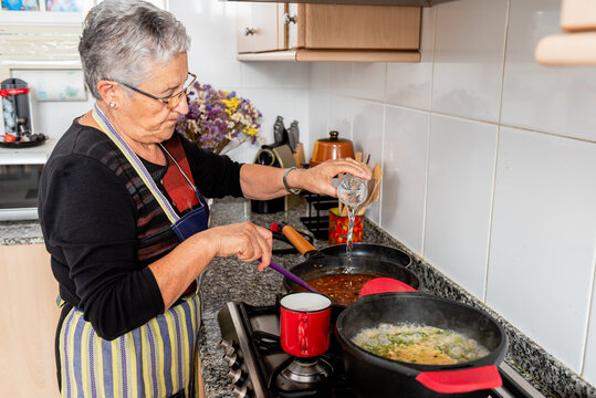Side view of senior housewife pouring water into frying pan with mixed ingredients while preparing traditional Catalan fish dish in home kitchen