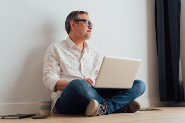 Contemplative senior male sitting with legs crossed on parquet with netbook on laps and cup of takeaway coffee in hand while looking away in thoughts