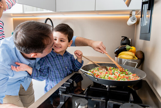 Happy young father and cute son sauteing cut ripe vegetables in frying pan while cooking lunch together in contemporary kitchen
