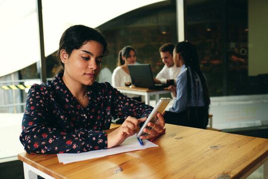 Young ethnic female text messaging on cellphone while sitting at table with paper sheets and pen