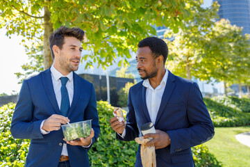 Positive young diverse coworkers in formal clothes smiling while having lunch break together on street on sunny day
