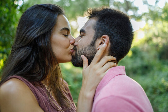 Side view young ethnic couple in casual outfits standing in lush green forest and kissing tenderly on sunny day