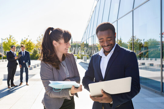 Focused young female manager in suit showing documents to African American male colleague standing near contemporary building with laptop on sunny day