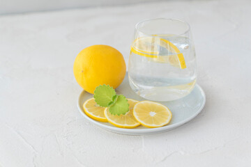transparent glass with pure water, lemon slices, mint leaf ,  whole lemon lie on white plate on light background. Healthy vitamin drink