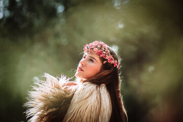 Side view of charming romantic young long haired female with pink floral wreath standing in forest looking away