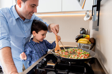Happy young father and cute son sauteing cut ripe vegetables in frying pan while cooking lunch together in contemporary kitchen