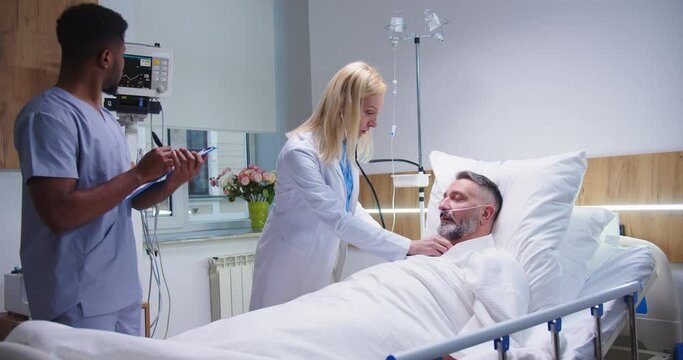 Caucasian pretty female professional doctor listen heartbeat of middle-aged miserable man patient using stethoscope in hospital while young African American male nurse assistant writing down symptoms