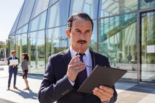 Serious middle aged male manager with mustache in classy outfit holding documents in hand and looking away while thinking about business strategy on street