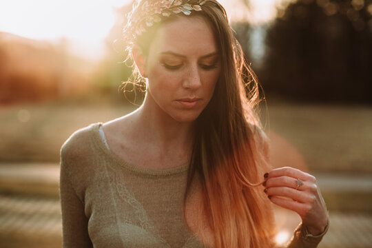 Crop serene female wearing transparent sweater standing in lush green park in evening sunlight and looking down pensively