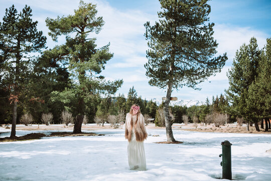 Young blonde female in boho style wedding dress and fur jacket with floral wreath on head standing on snowy meadow against forest