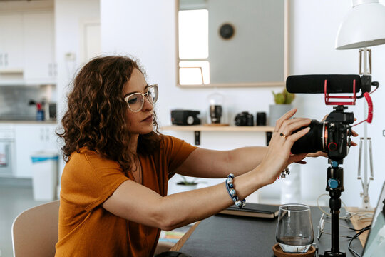 Side view of focused young female blogger with curly hair in casual outfit and eyeglasses setting professional photo camera placed on tripod before shooting video at home