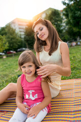 woman mom, straightens her hair for child, little girl 5-7 years old, laughs, smiles, joyful cheerful, outdoor recreation on weekend. They sit on a blanket in summer. Trees and green grass background.