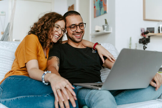 Cheerful young diverse couple in casual clothes and eyeglasses holding hands while sitting together on sofa and having video call on laptop