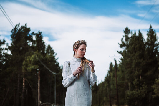 Charming young female in white casual outfit and hairband touching hair gently while standing near coniferous woods and looking at camera