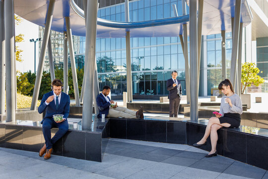 Group of young multiracial coworkers in classy outfits sitting on bench near modern business center and having lunch
