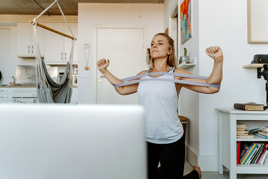 Sportswoman performing arms exercise with resistance bands while watching tutorials on laptop and working out at home