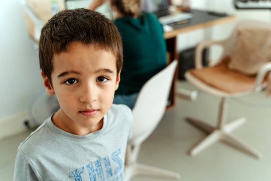 From above content schoolboy in casual shirt standing in light room near desk with computer and looking at camera