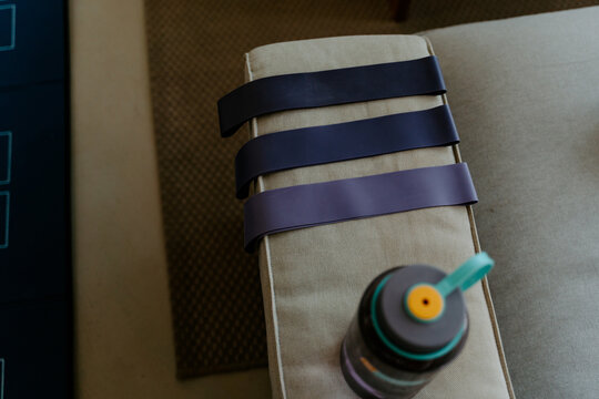 High angle collection of elastic bands of different resistance levels placed on cozy couch arm near water bottle before exercising