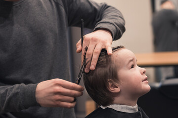 Obraz na płótnie Canvas A pretty boy toddler happy to be on the haircut with a professional children's hairdresser. Blond little boy having a haircut at hair salon. Hairdresser's hands making hairstyle to child.