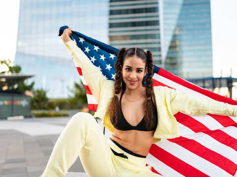 Side view of confident young ethnic lady in sports bra waving American flag and looking at camera against modern glass skyscraper