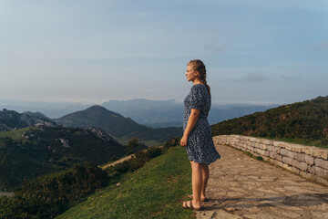 Side view full body young positive female impressed by amazing mountainous landscape standing on green hill and admiring views