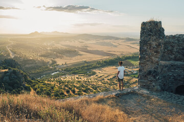 Back view of distant faceless male with long curly hair wearing white shirt standing on sunny hilltop and admiring vast scenic valley with tranquil pond and lush greenery