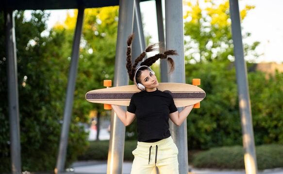 Happy young ethnic female with curly hair in activewear holding longboard behind back and listening to music in headphones during training in skate park