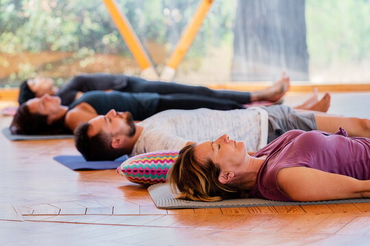 Side view of people with closed eyes lying on mats after practicing yoga on wooden floor in class