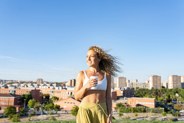 Cheerful young female wearing white sports bra standing with takeaway coffee against modern city buildings on sunny day while looking away with toothy smile